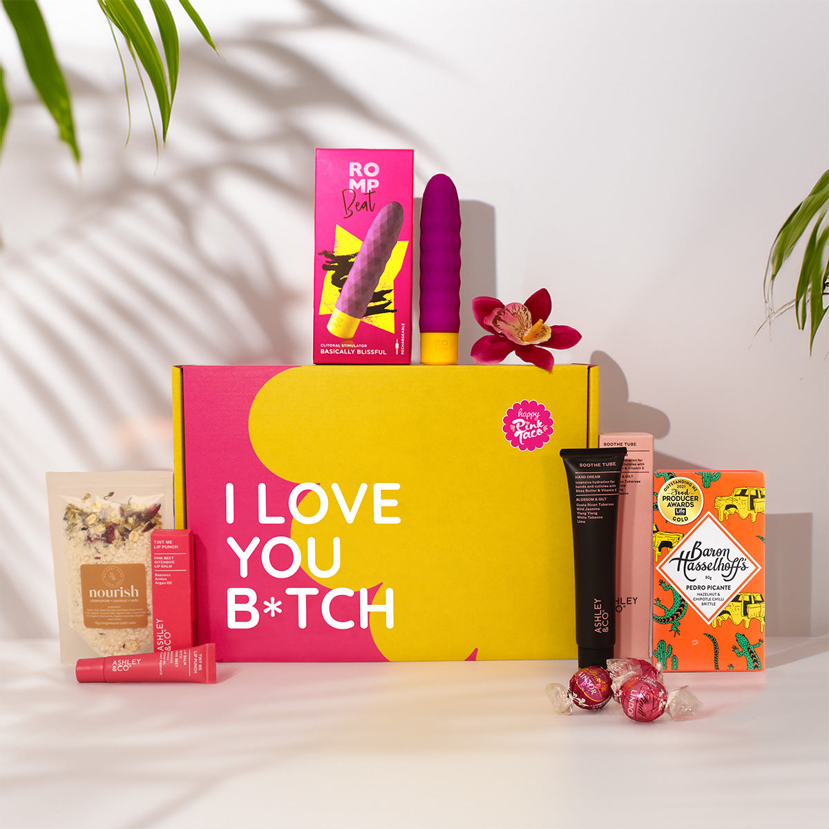 A curated gift box set on a white surface with leaf shadows. The main item is a large pink and yellow box reading 'I LOVE YOU B*TCH' in white text.