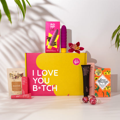 A curated gift box set on a white surface with leaf shadows. The main item is a large pink and yellow box reading 'I LOVE YOU B*TCH' in white text.