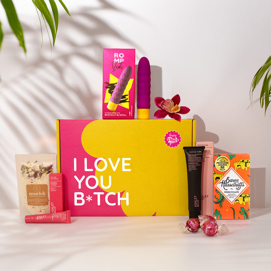 A curated gift box set on a white surface with leaf shadows. The main item is a large pink and yellow box reading 'I LOVE YOU B*TCH' in white text.