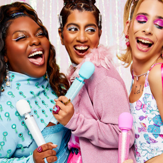 Three women with colorful hair and outfits, holding toy Le Wand Massagers against a light background.