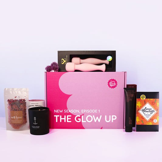 A curated beauty and self-care gift set arranged on a white surface against a pale lavender background. The set includes a large pink box labeled 'NEW SEASON, EPISODE 1 THE GLOW UP,' two pink silicone personal massagers in a black box behind it, a black candle jar with an anchor logo, a pouch of dried botanicals labeled 'self love,' a black tube labeled 'glow,' and a square package labeled 'Bloom Naturally.
