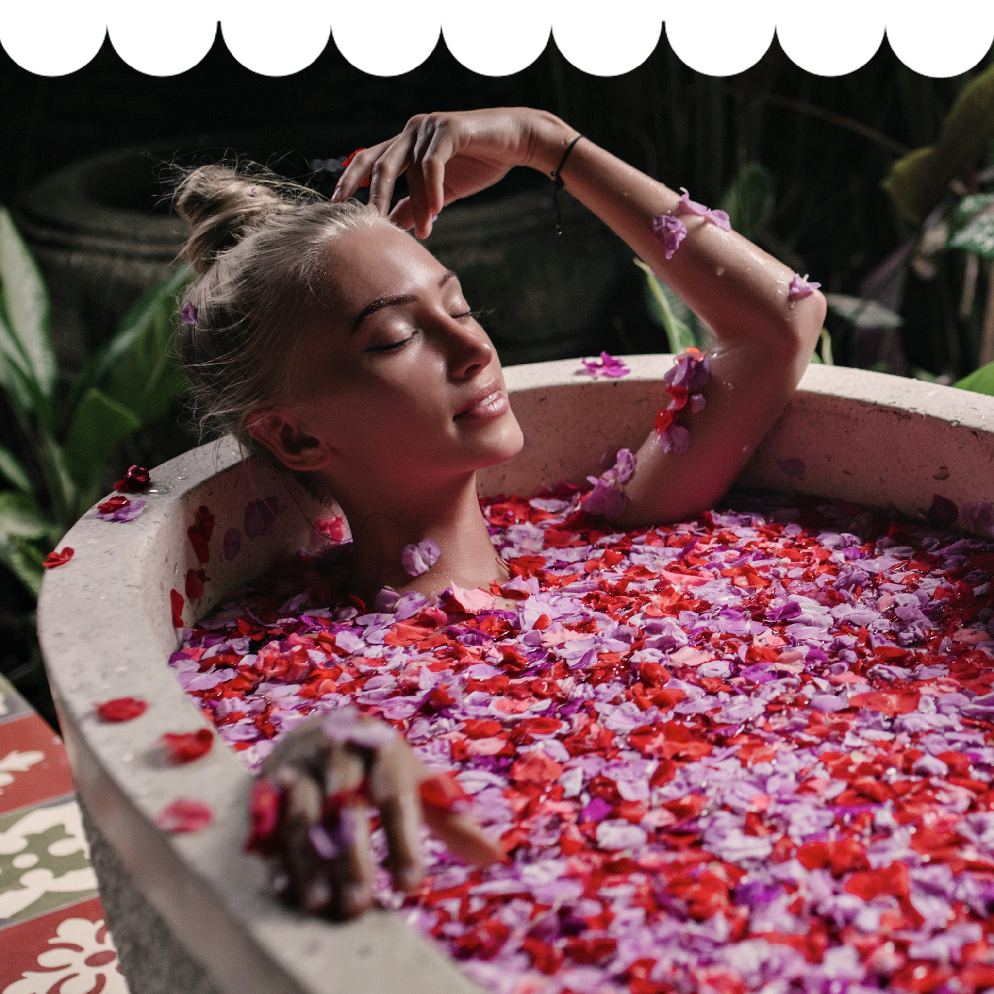 Woman indulging in Self-care, relaxing in a bathtub filled with red and pink flower petals surrounded by greenery.