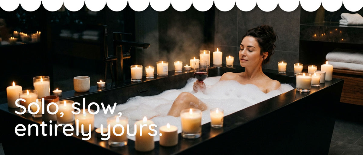 A woman relaxes in a bubble bath surrounded by lit candles in a dark spa-like setting. Large overlaid text reads 'Solo, slow, entirely yours.